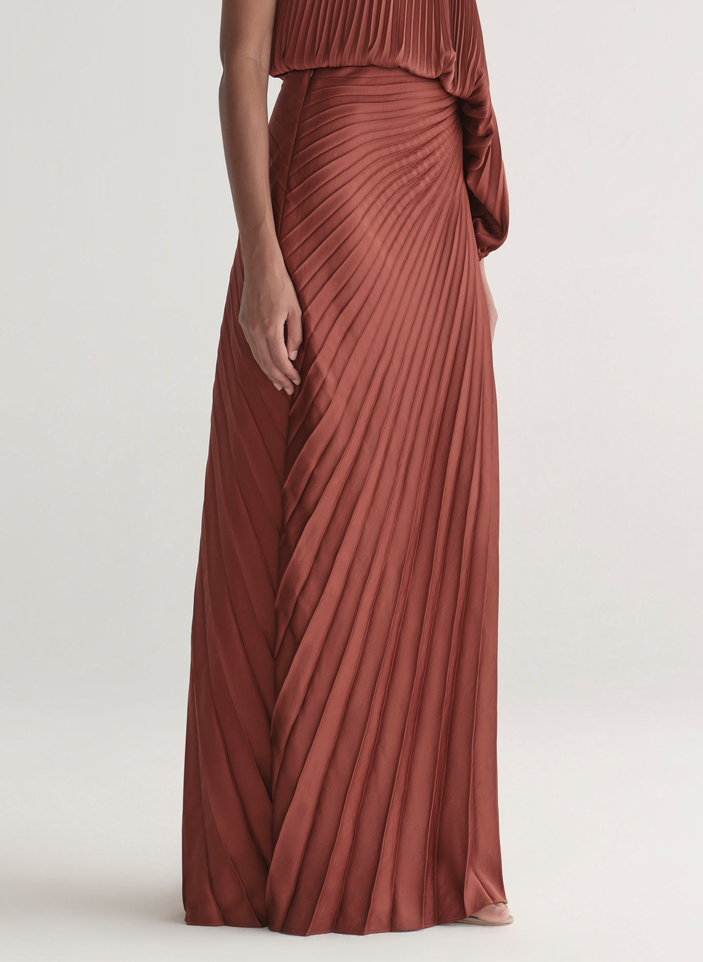 Woman wearing a one-shoulder rust-colored top with matching skirt with pleated details on a white background