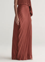 Woman wearing a one-shoulder rust-colored top with matching skirt with pleated details on a white background