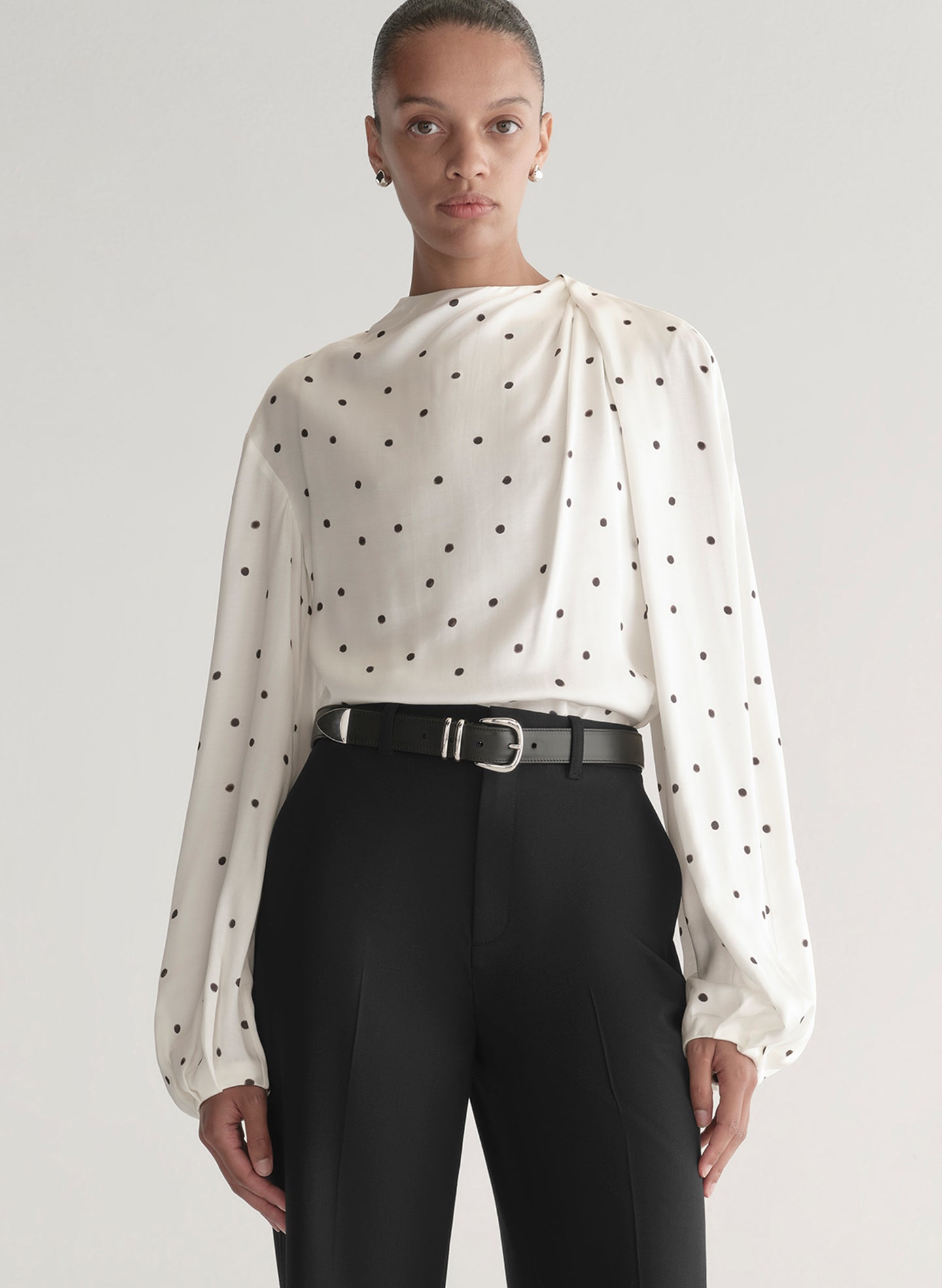 Woman wearing a white blouse with black polka dots and black pants on a plain background
