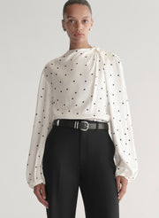 Woman wearing a white blouse with black polka dots and black pants on a plain background