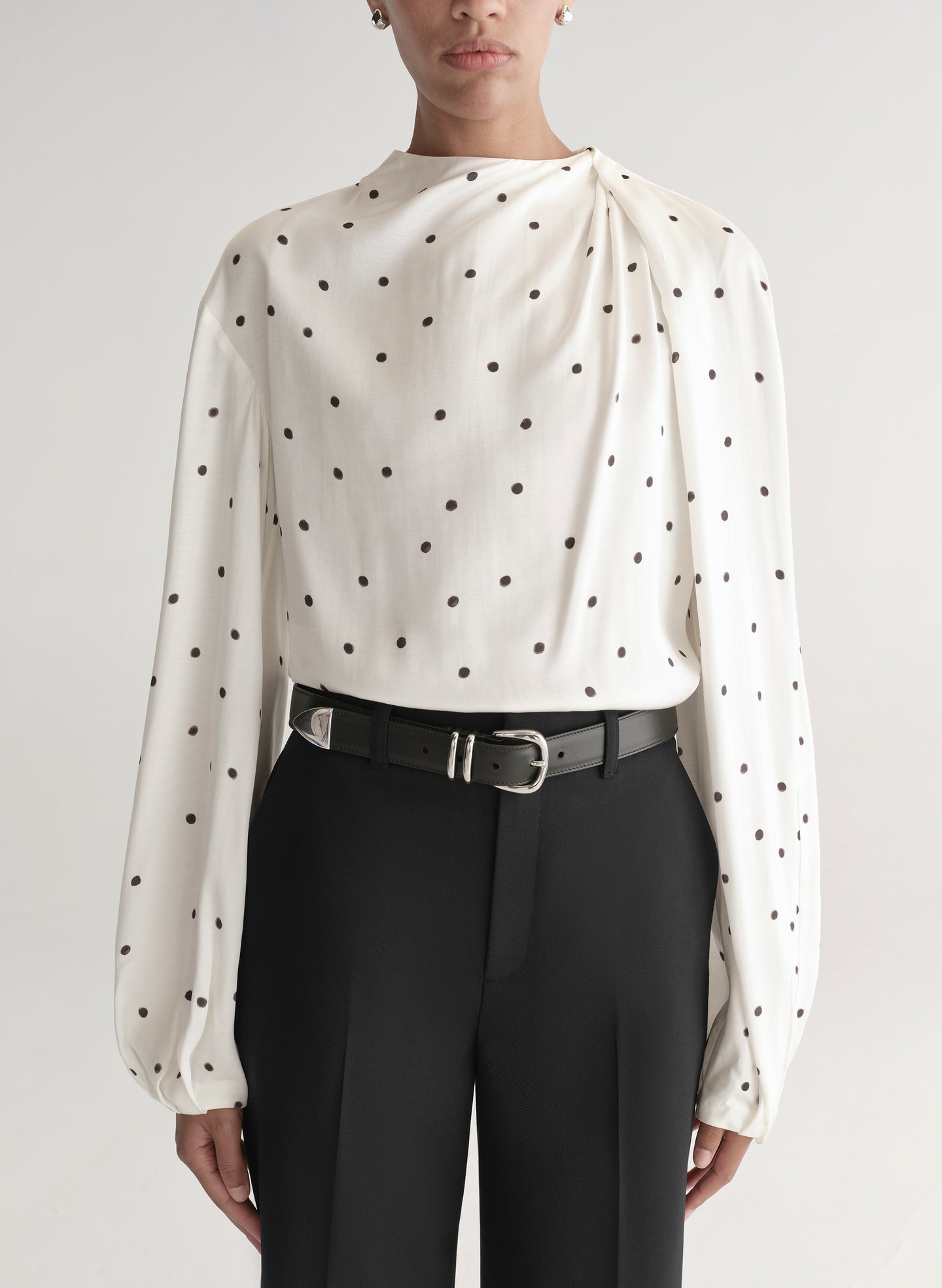 Woman wearing a white blouse with black polka dots and black pants on a plain background