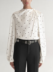 Woman wearing a white blouse with black polka dots and black pants on a plain background