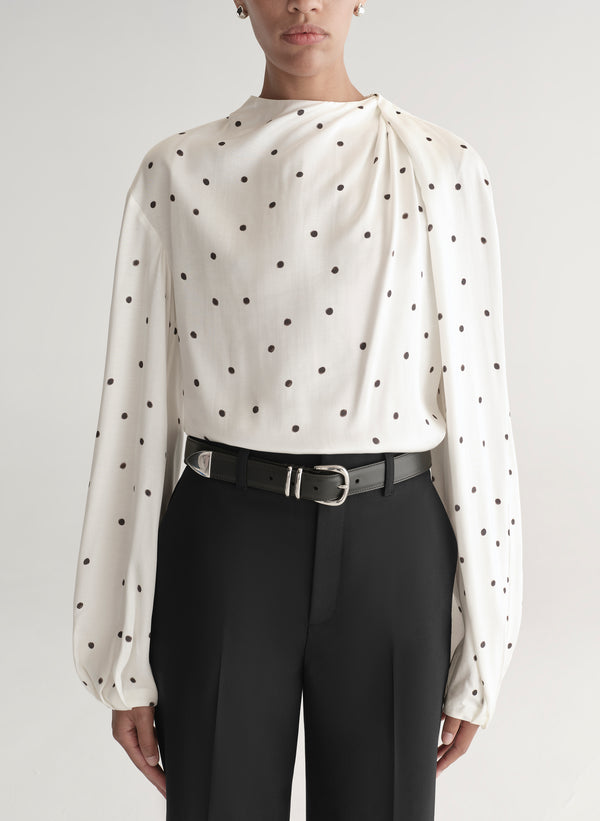 Woman wearing a white blouse with black polka dots and black pants on a plain background