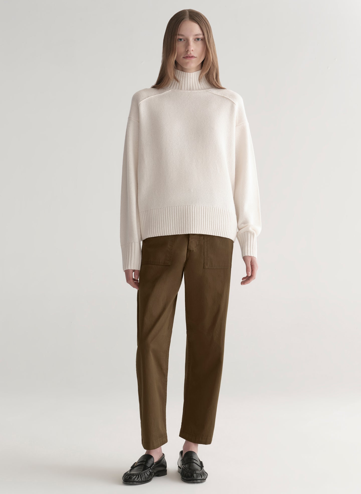 Woman wearing a white sweater and olive green pants on a plain background