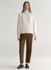 Woman wearing a white sweater and olive green pants on a plain background