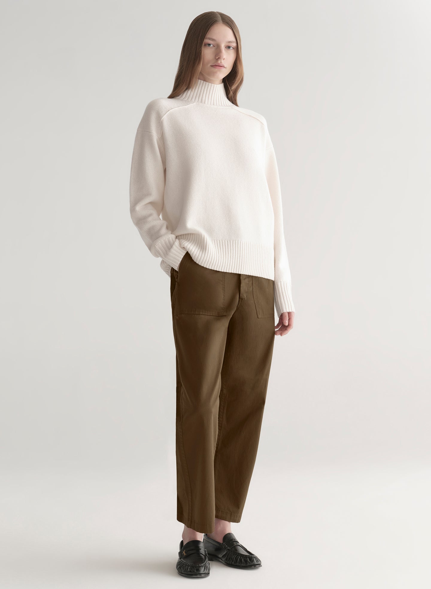 Woman wearing a white sweater and olive green pants on a plain background