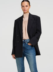 A model wearing a black double-breasted blazer over a beige pleated top and blue jeans, photographed against a light gray studio background.