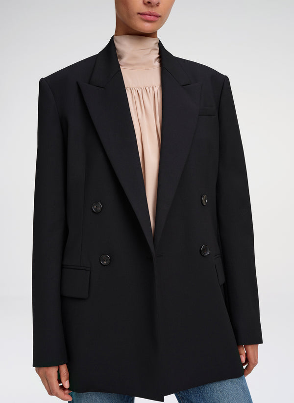 A model wearing a black double-breasted blazer over a beige high-neck blouse and blue jeans, shown from the neck down against a light gray studio background.