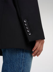 Close-up of a person wearing a black blazer with five dark buttons on the sleeve cuff, paired with blue jeans, against a light gray background.