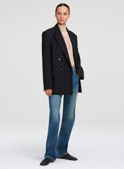 A model stands against a light gray studio background wearing an oversized black double-breasted blazer over a light beige turtleneck, blue flare jeans, and black loafers.