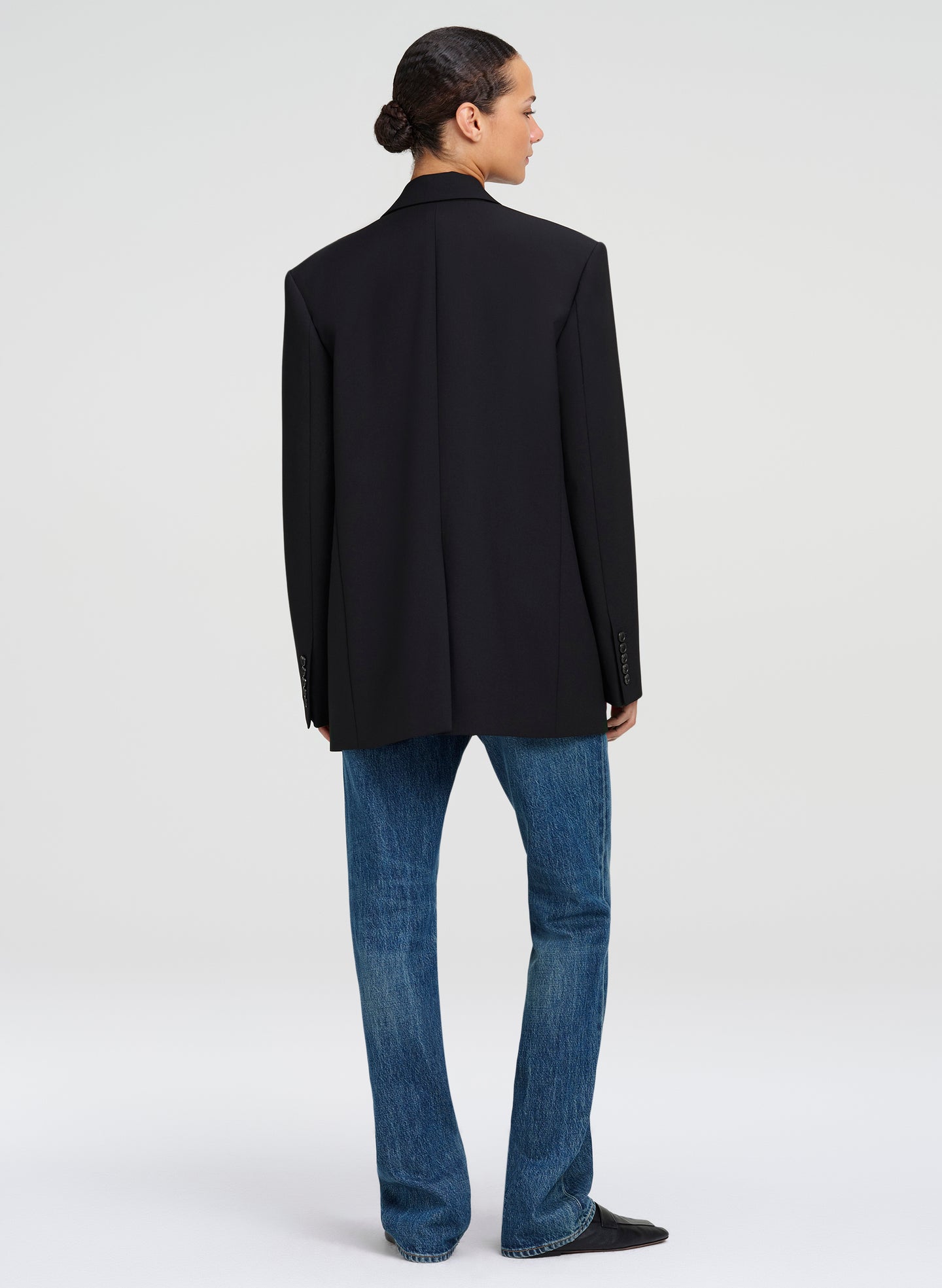 A man wearing a black blazer and blue jeans, photographed from the back against a light gray studio background.