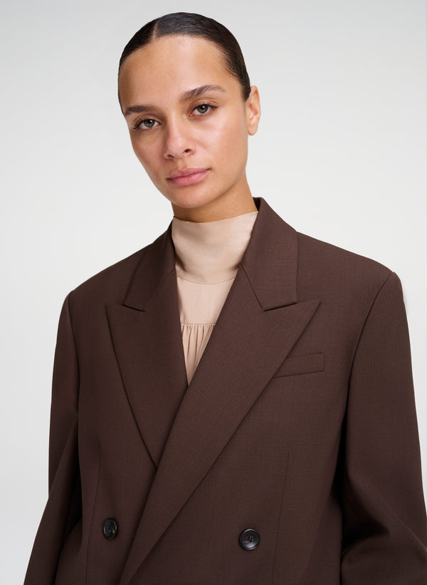 A close-up of a model wearing a dark brown double-breasted blazer over a beige mock neck top, shot against a light gray studio background.