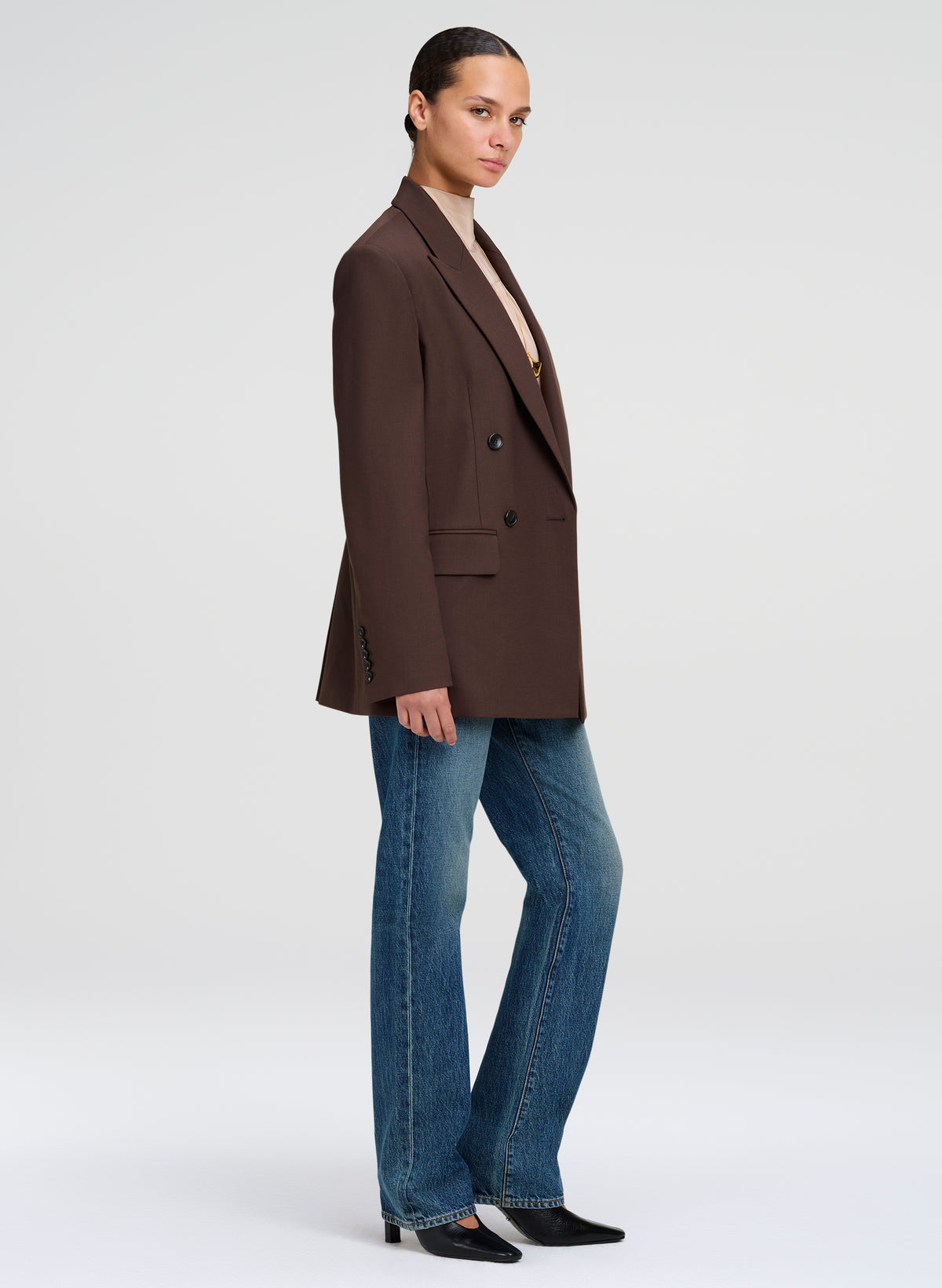 A model wearing a dark brown double-breasted blazer, blue jeans, and black shoes, shown in a side profile against a light gray studio background.