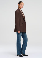 A model wearing a dark brown double-breasted blazer, blue jeans, and black shoes, shown in a side profile against a light gray studio background.