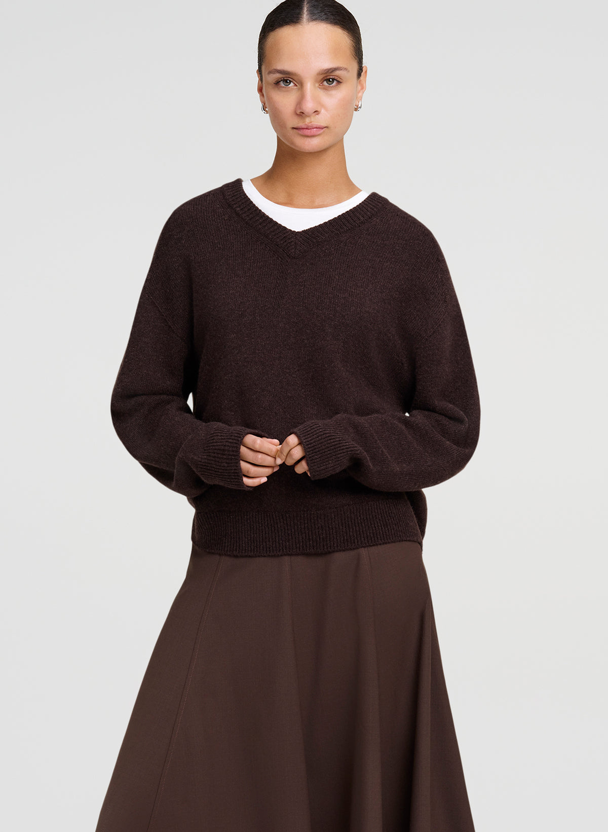 A woman modeling a dark brown knit sweater over a white undershirt, paired with a matching brown skirt, against a light gray studio background.