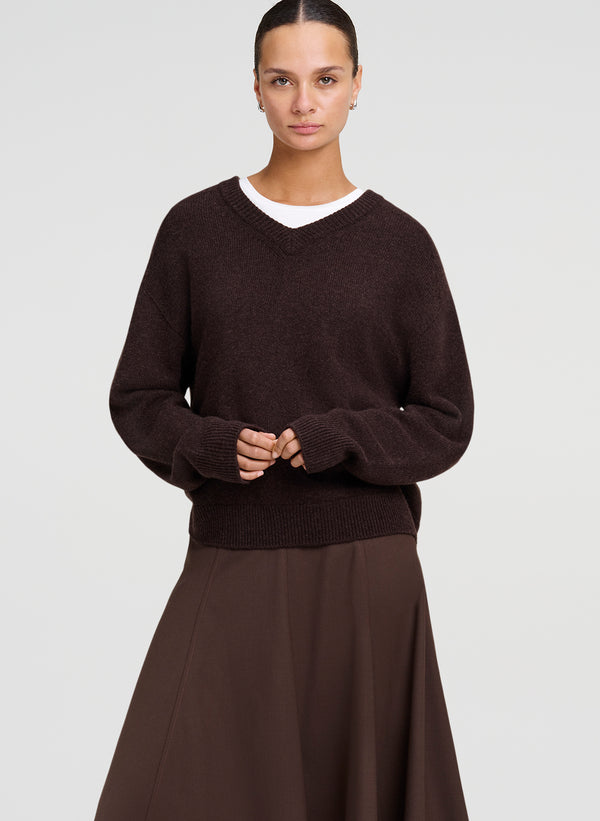 A woman modeling a dark brown knit sweater over a white undershirt, paired with a matching brown skirt, against a light gray studio background.