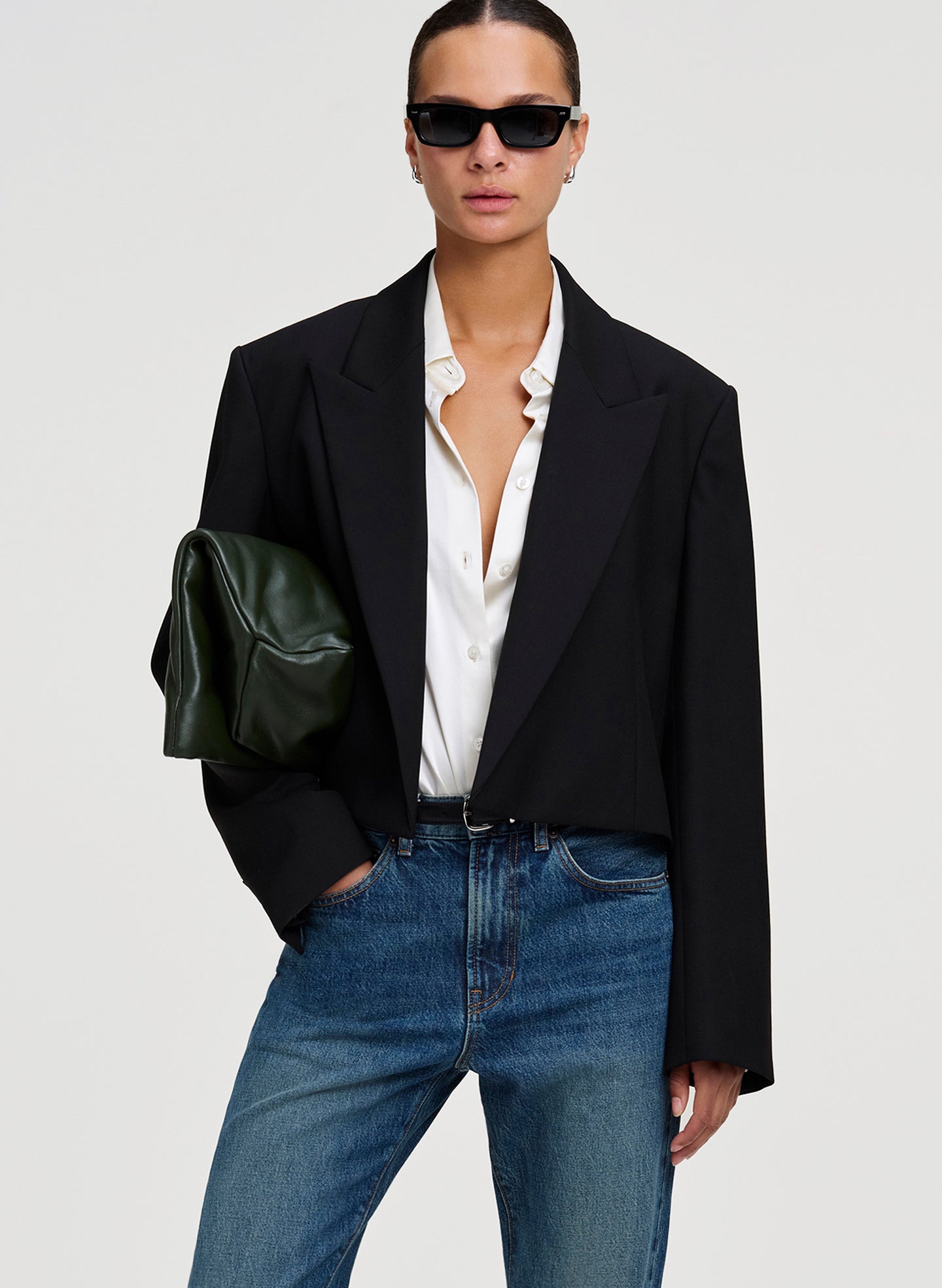 A model wearing a black oversized blazer, white button-down shirt, blue jeans, and black sunglasses, holding a dark green leather clutch against a light gray studio background.