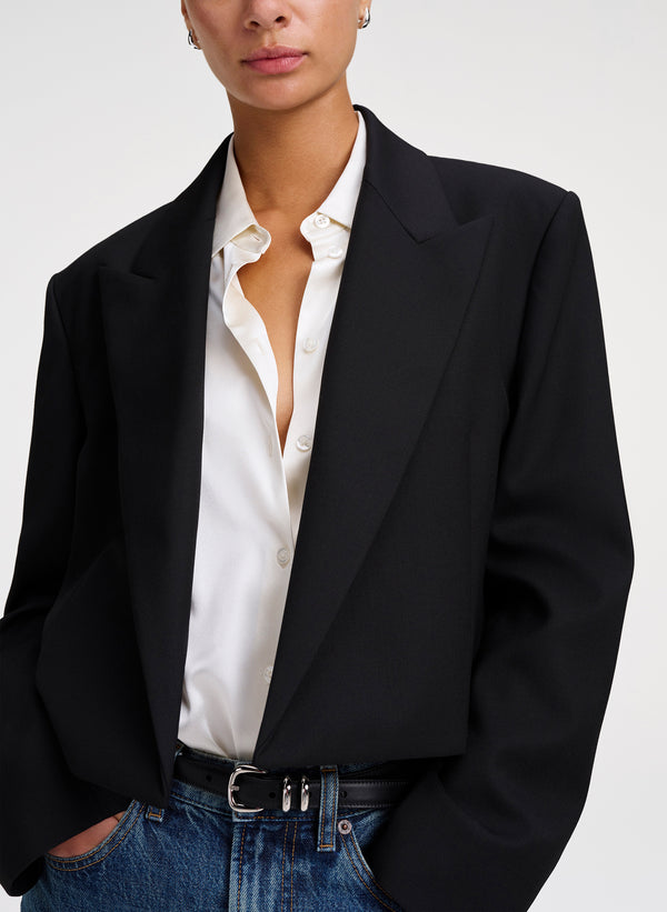 A close-up of a model wearing an oversized black blazer over a white button-down shirt and blue jeans, photographed against a light gray studio background.