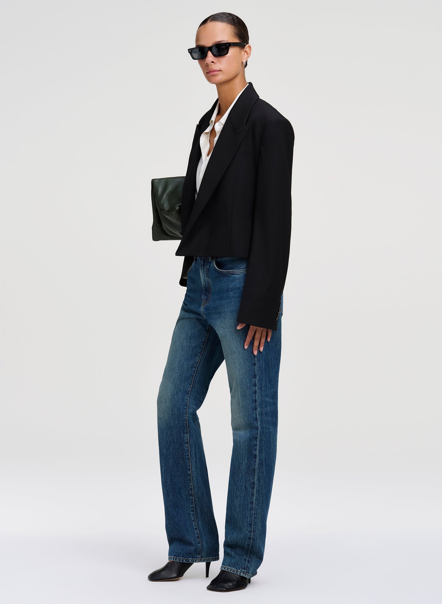 A model stands in profile wearing a black cropped blazer, blue straight-leg jeans, black pointed heels, and black sunglasses, holding a small dark green clutch against a clean light gray studio background.