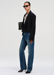 A model stands in profile wearing a black cropped blazer, blue straight-leg jeans, black pointed heels, and black sunglasses, holding a small dark green clutch against a clean light gray studio background.