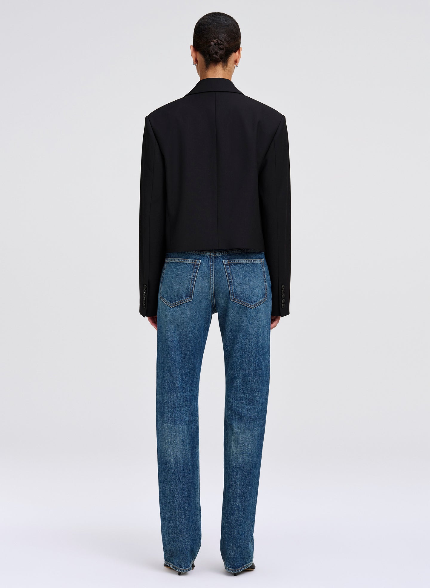 A model shown from the back wearing a black cropped blazer and blue jeans against a light gray studio background.