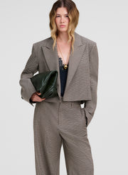 A model wearing a cropped taupe-gray blazer and matching wide-leg trousers, holding a dark green leather clutch against a light gray studio background.