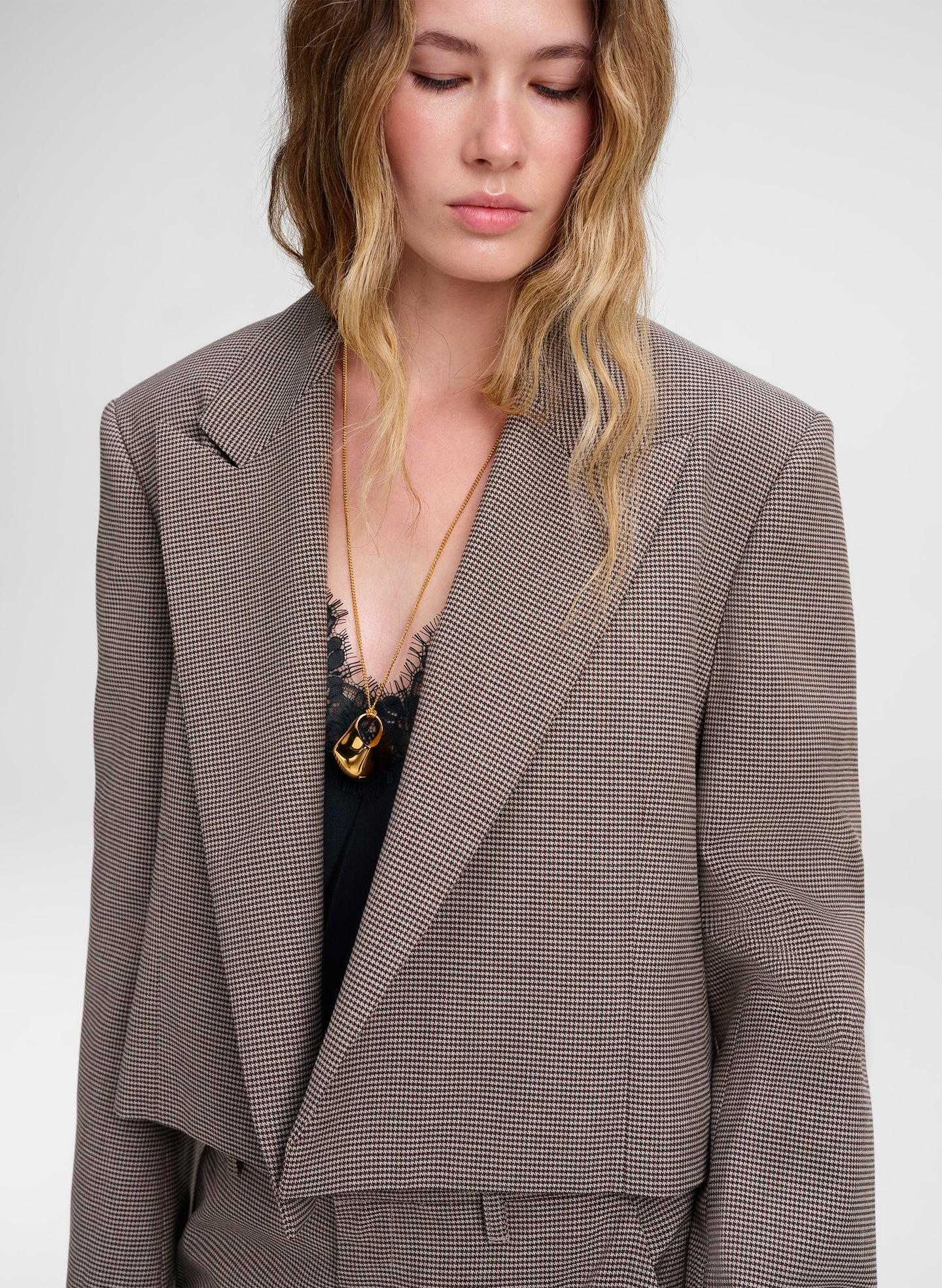 A woman wearing a cropped, structured blazer in a fine brown and beige houndstooth pattern, styled with a black top and a gold necklace.