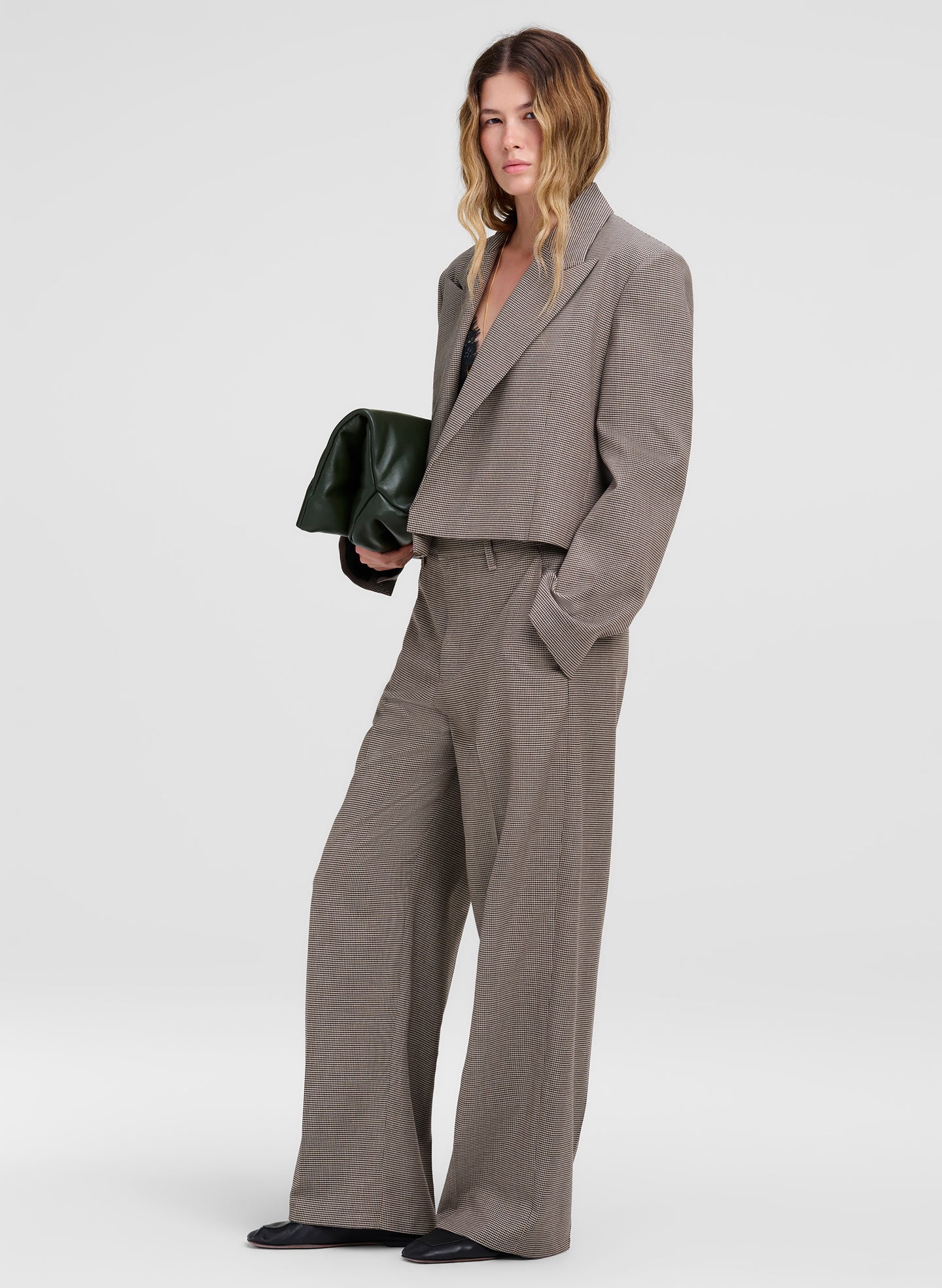 A model wearing a matching taupe gray cropped blazer and wide-leg trousers suit, holding a dark green clutch against a light gray studio background.