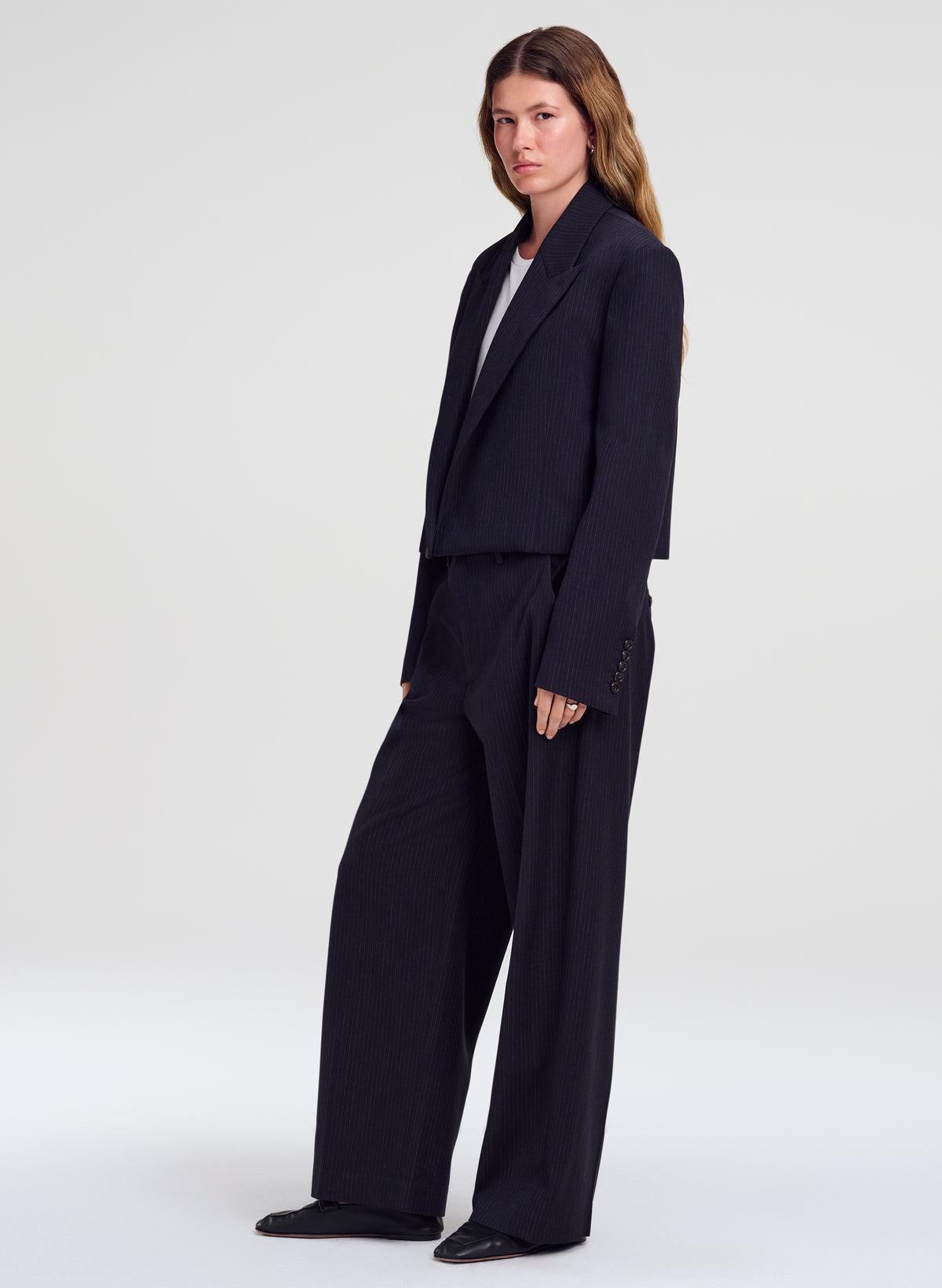 A woman modeling a dark navy blue tailored suit consisting of a cropped blazer and wide-leg trousers, styled over a white top, against a light gray studio background.
