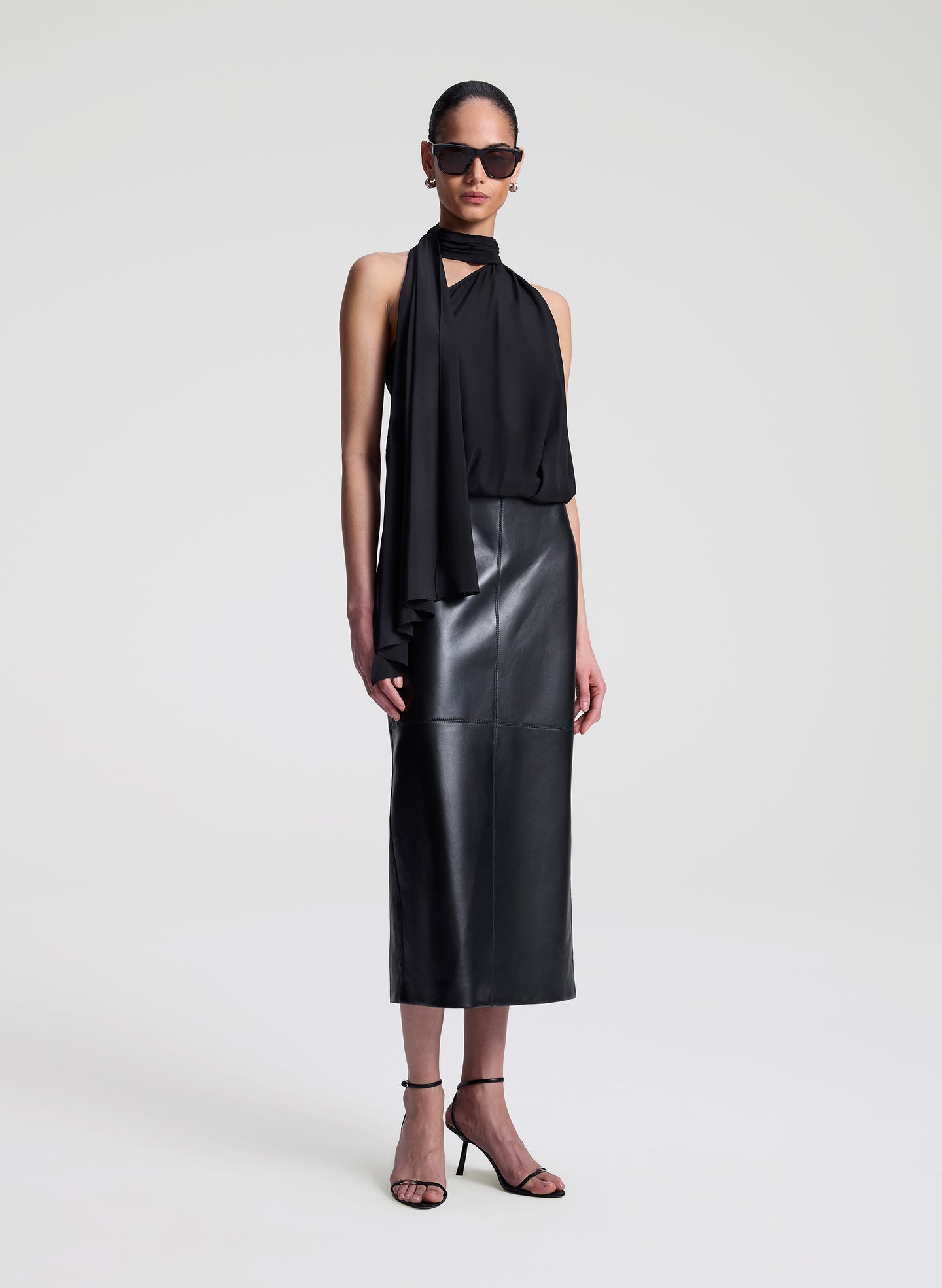 Woman wearing a black sleeveless top with a draped design and a leather skirt on a white background