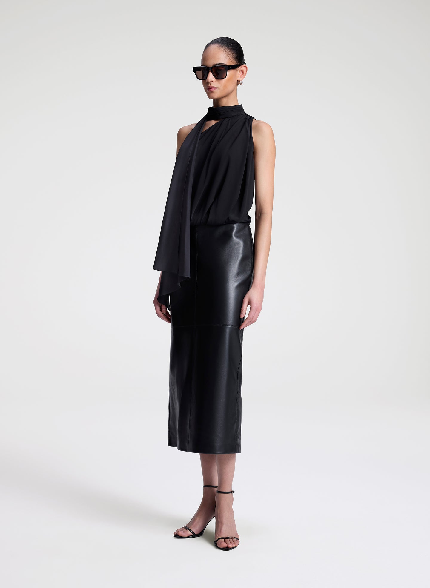 Woman wearing a black sleeveless top with a draped design and a leather skirt on a white background
