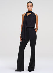 A model wearing a black sleeveless halter-neck top with a draped asymmetrical overlay and wide-leg pants, photographed against a light gray studio background.