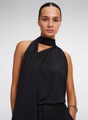 A woman modeling a black halter-neck top with a draped, asymmetrical shawl collar and side cutout, paired with black trousers against a light gray studio background.