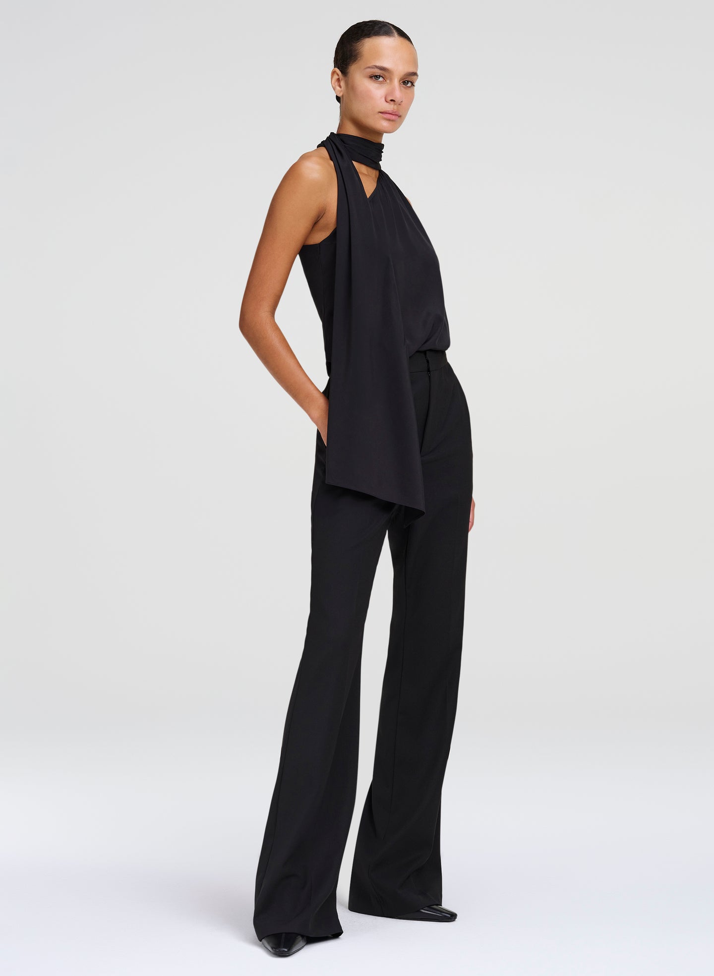 A model wearing a black sleeveless high-neck top with draped fabric details and wide-leg pants, photographed against a light gray studio background.