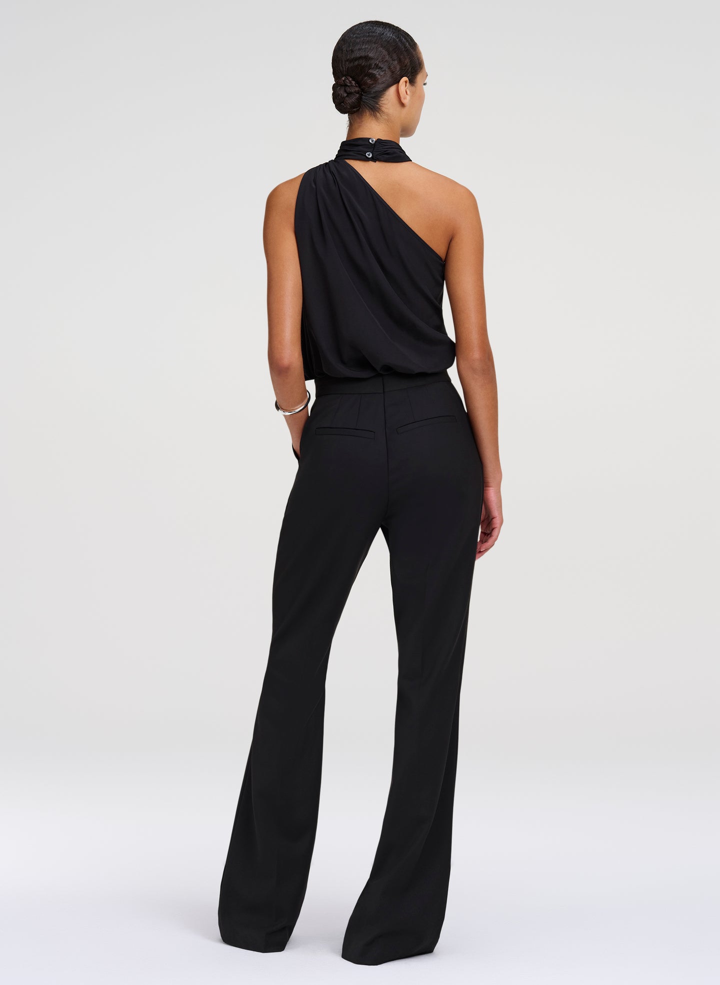 A model shown from the back wearing a black one-shoulder draped top and matching black wide-leg trousers, photographed against a light gray studio background.