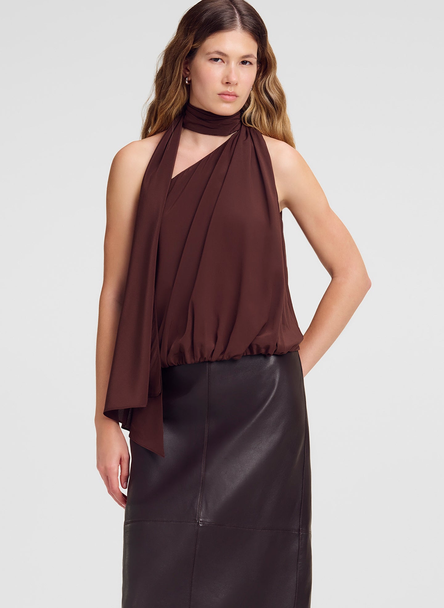 A woman modeling a sleeveless halter-neck top in dark brown with draped fabric details, paired with a dark skirt, against a light gray studio background.