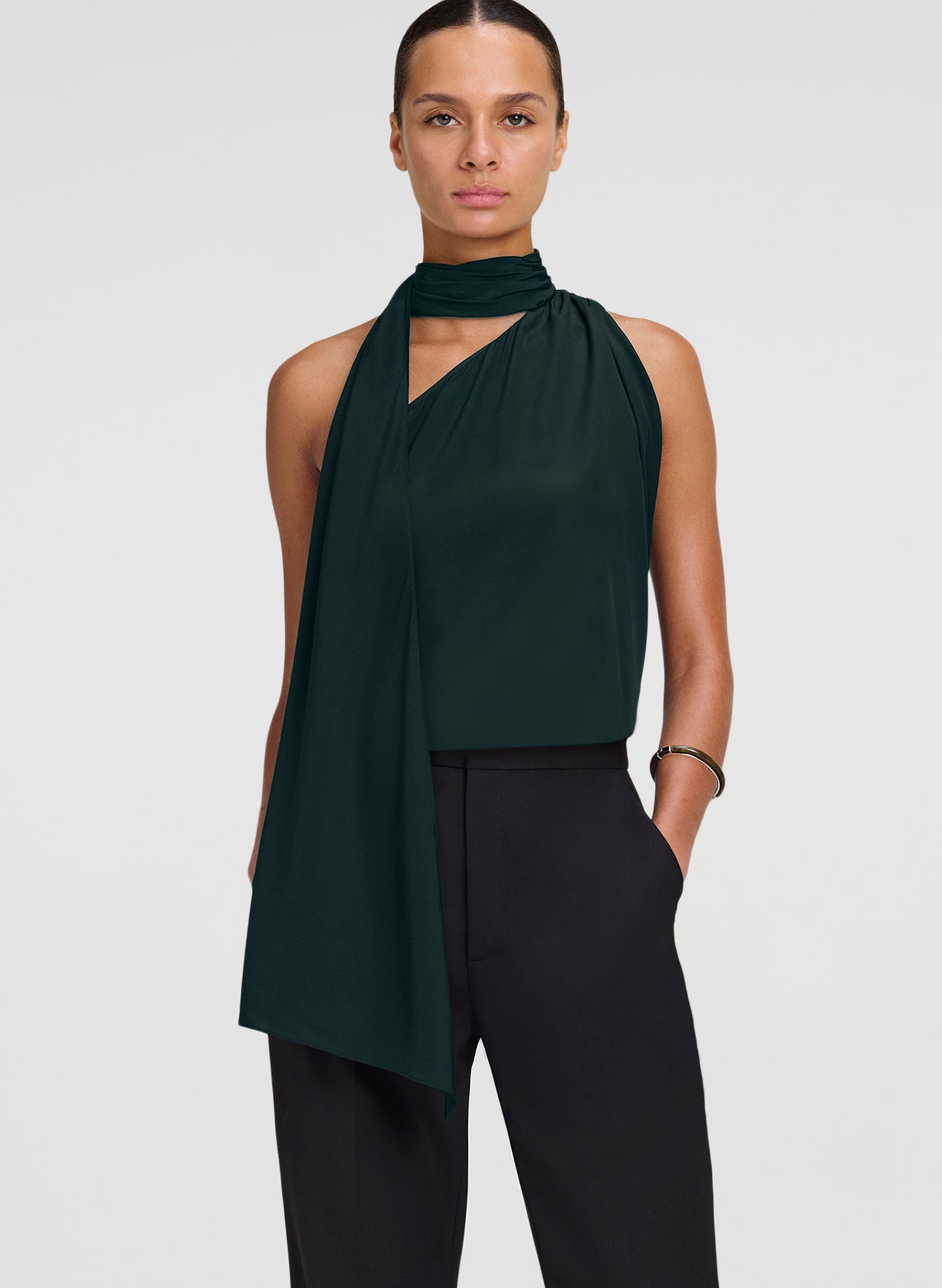 A model wearing a dark forest green halter top with a draped asymmetrical scarf detail and black trousers, photographed against a solid light gray background.