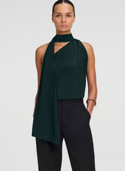 A model wearing a dark forest green halter top with a draped asymmetrical scarf detail and black trousers, photographed against a solid light gray background.