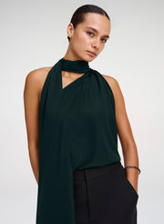 A model wearing a dark forest green halter-neck top with a draped scarf detail and black trousers, photographed against a light gray studio background.