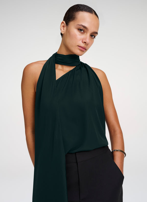 A model wearing a dark forest green halter-neck top with a draped scarf detail and black trousers, photographed against a light gray studio background.