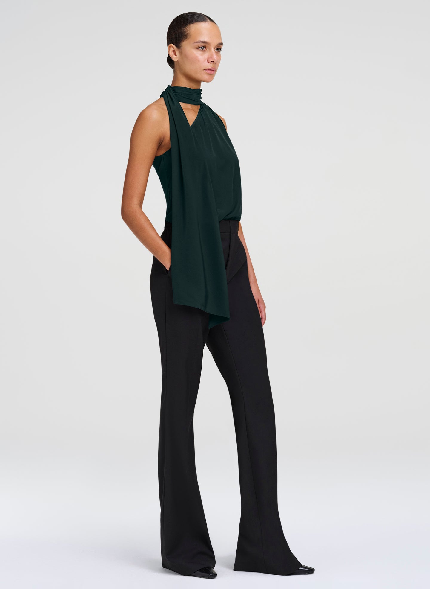 A model is shown in profile wearing a dark forest green sleeveless halter top with draped fabric and black wide-leg trousers, set against a plain light gray studio background.