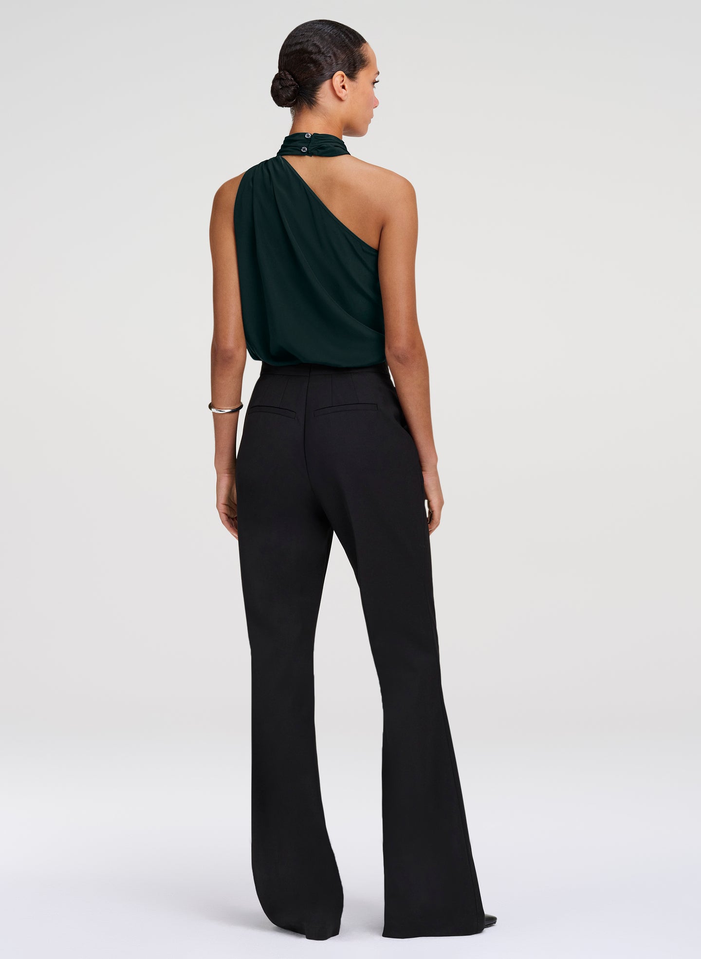 A model shown from the back wearing a dark green one-shoulder draped top and black flared pants against a light gray studio background.