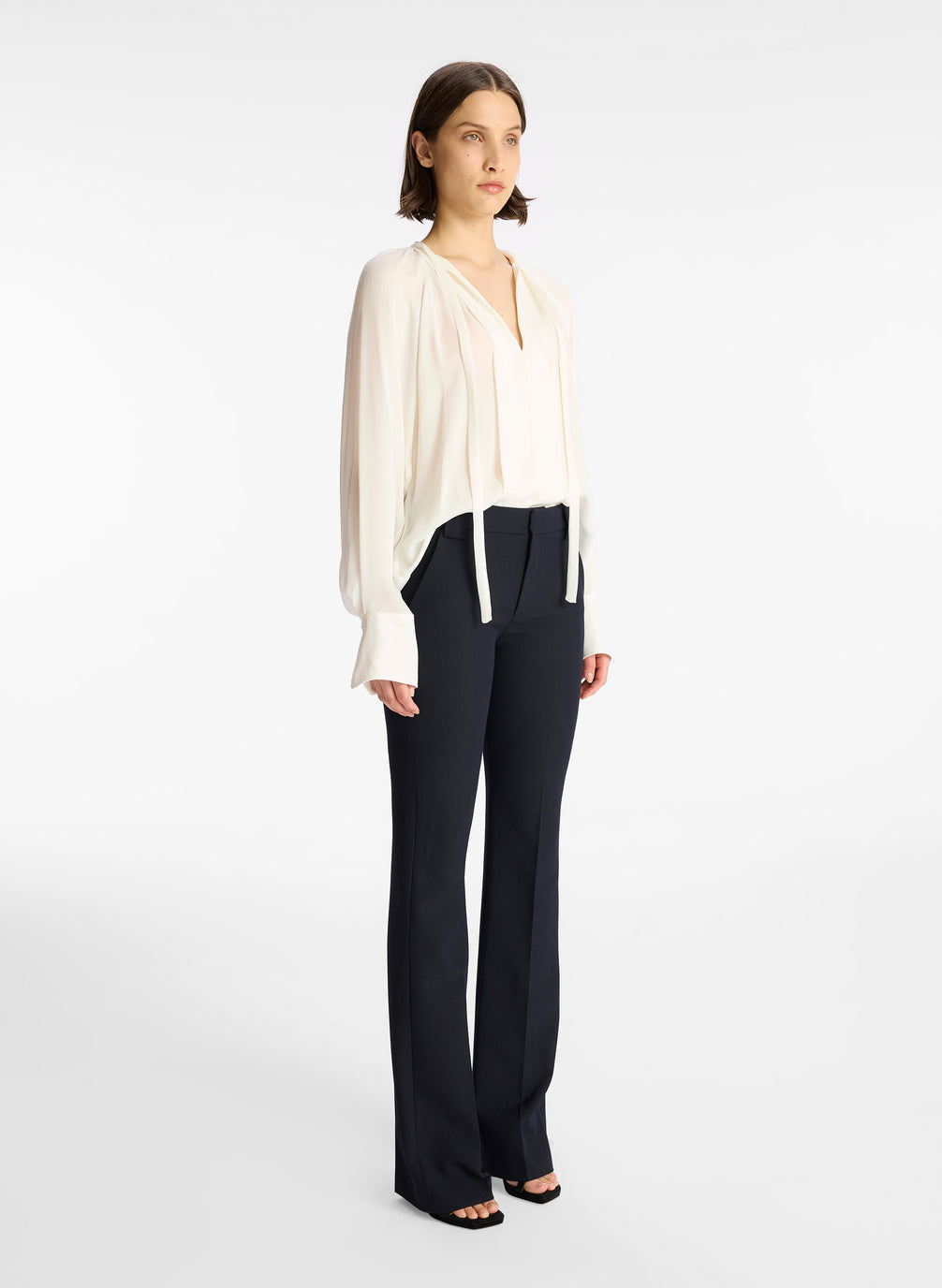 side view of woman wearing cream long sleeve v neck blouse and navy blue pants