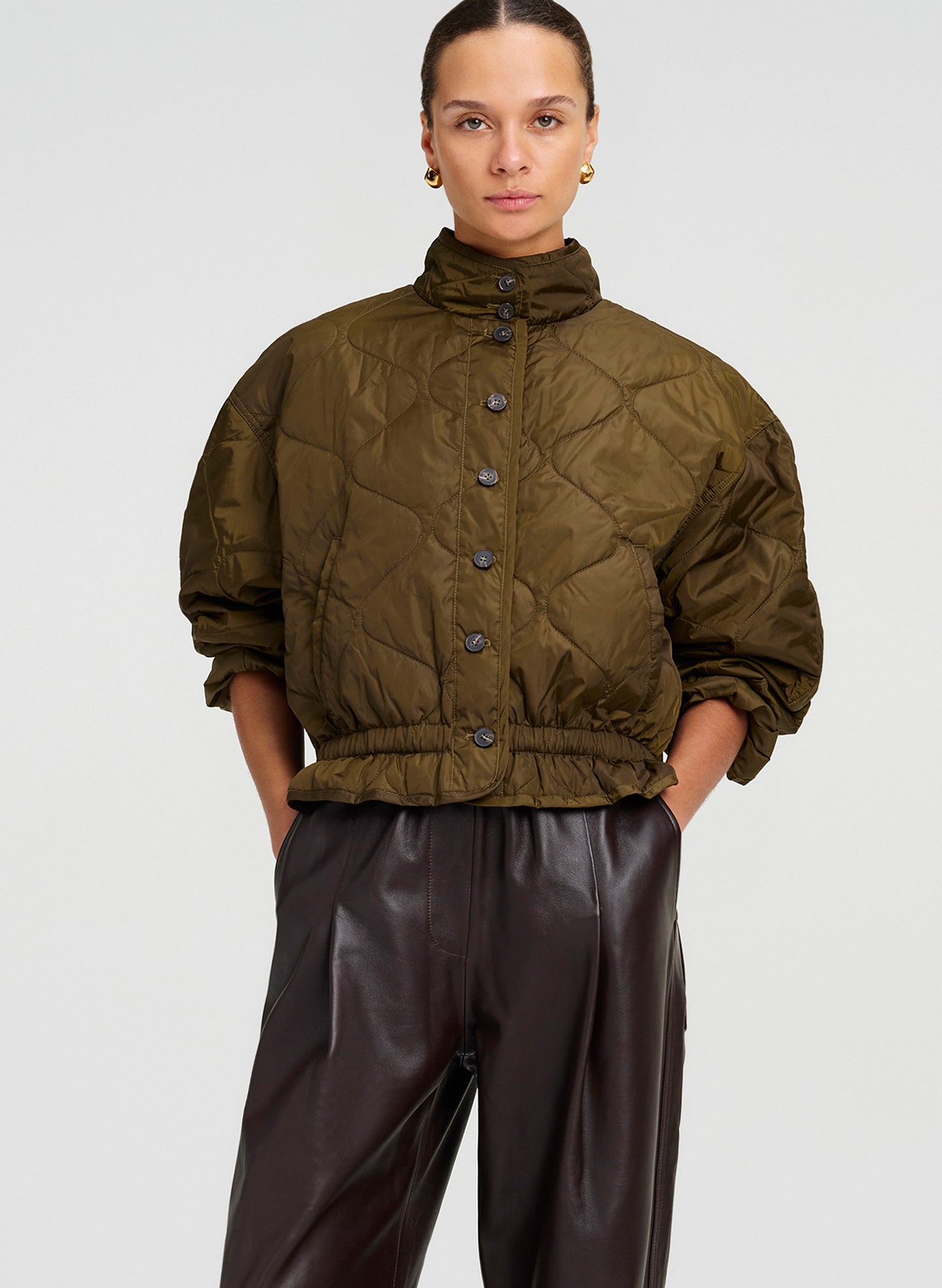 A model wearing an olive green quilted cropped jacket with a high collar and dark brown leather pants, photographed against a light gray studio background.