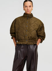 A model wearing an olive green quilted cropped jacket with a high collar and dark brown leather pants, photographed against a light gray studio background.