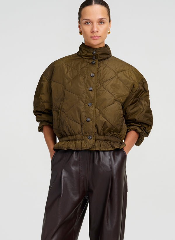 A model wearing an olive green quilted cropped jacket with a high collar and dark brown leather pants, photographed against a light gray studio background.