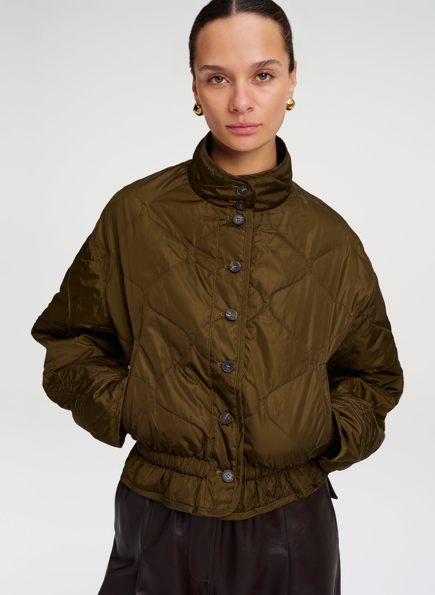 A model wearing an olive green quilted puffer jacket with a high stand-up collar and front button closure, paired with black pants, against a light gray studio background.