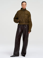 A model stands against a light gray studio background wearing an olive green quilted cropped jacket with a high mock neck and black wide-leg leather pants.