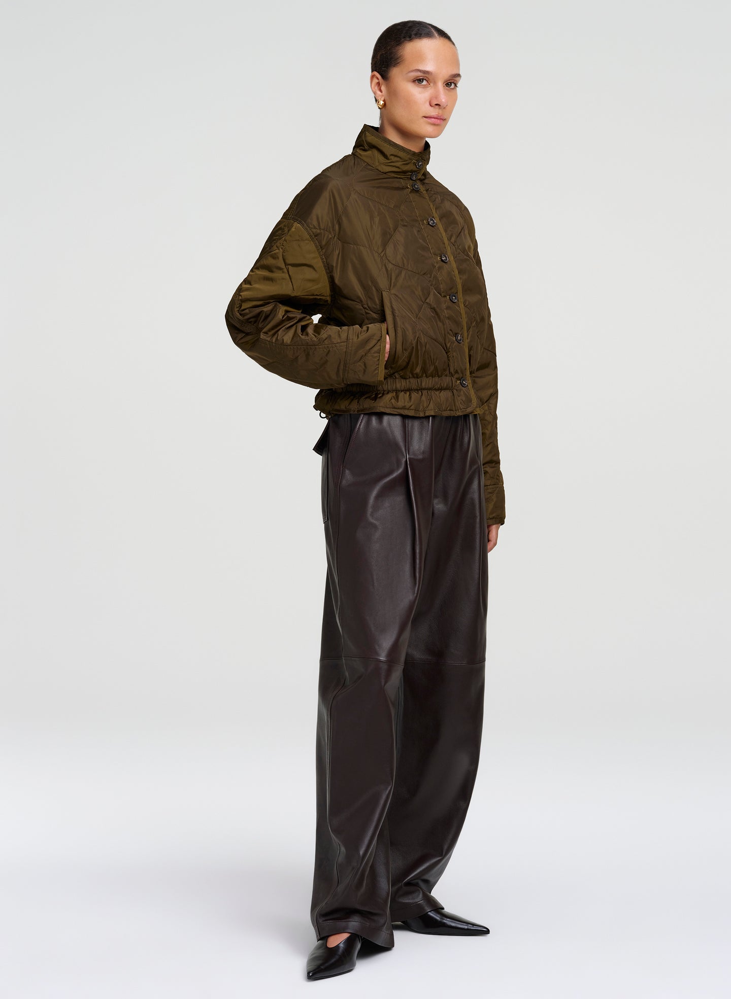A model stands in profile wearing a cropped quilted jacket in dark olive green and black wide-leg pants against a light gray studio background.