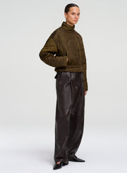 A model stands in profile wearing a cropped quilted jacket in dark olive green and black wide-leg pants against a light gray studio background.